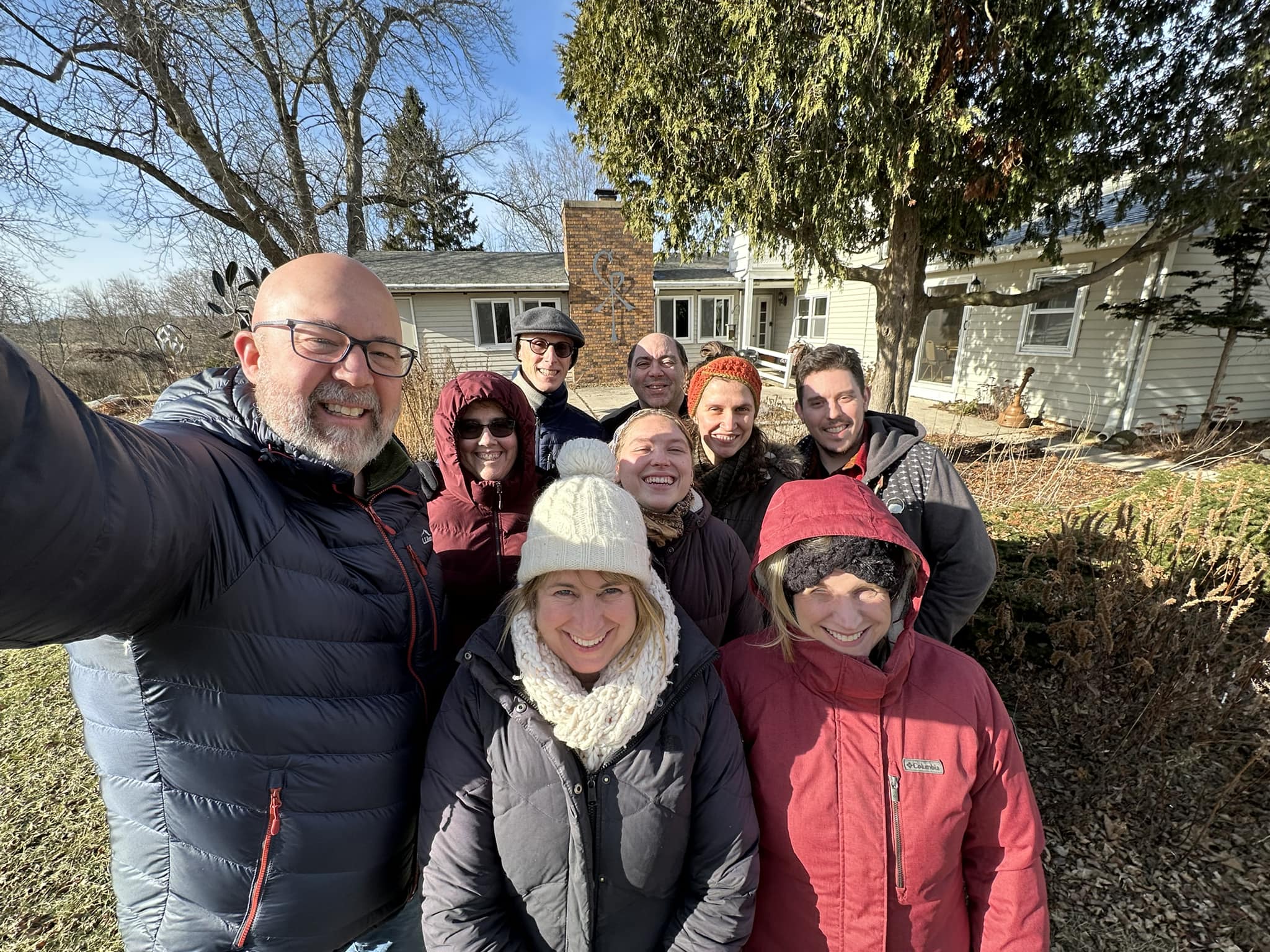 A group of nine people in winter clothing smile for a photo outside a house with trees and a chimney in the background.