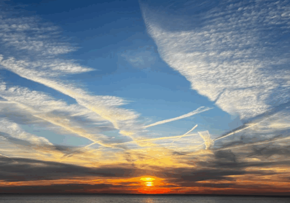 Sunset over ocean with vibrant orange and blue sky. Wispy clouds create dynamic patterns, reflecting light on calm water below. No landmarks visible.