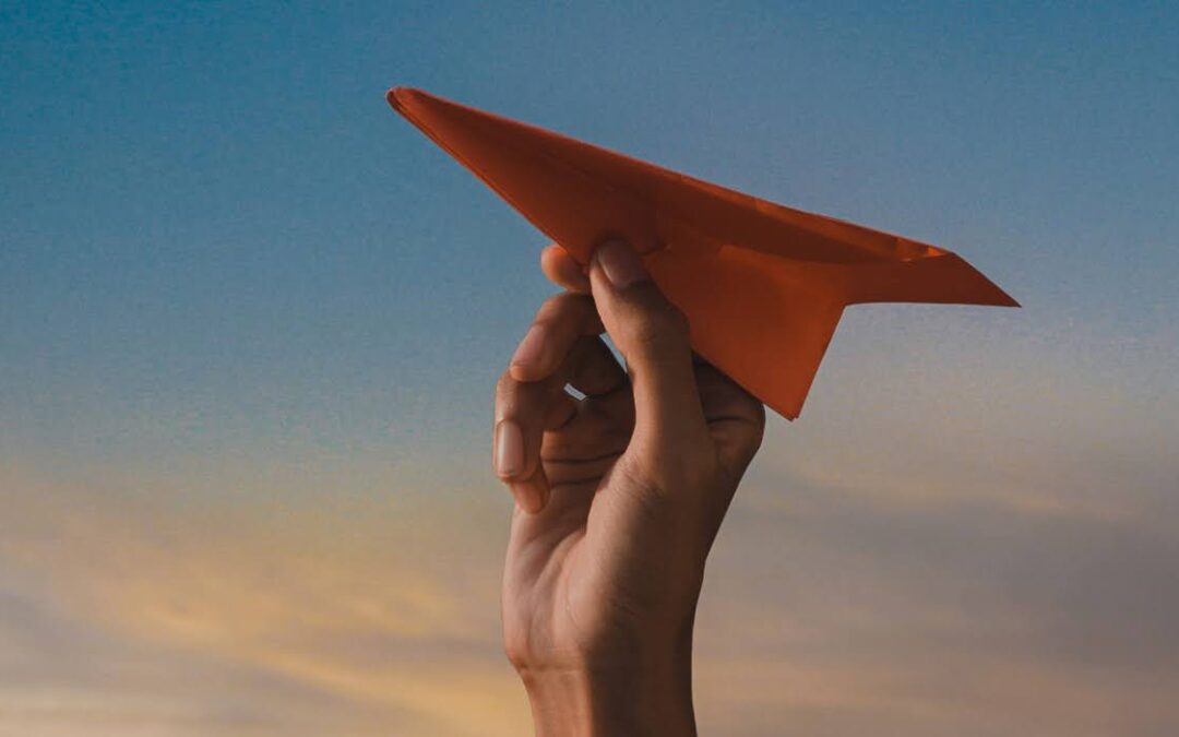 A person's hand holds an orange paper airplane against a backdrop of a dusky sky with shades of blue and orange.