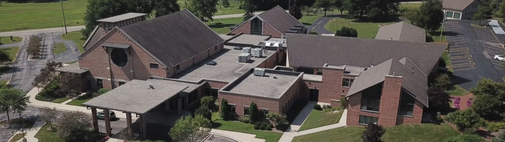 Aerial view of a large brick building complex with sloped roofs, surrounded by greenery and parking areas. Well-maintained landscape and roads visible.