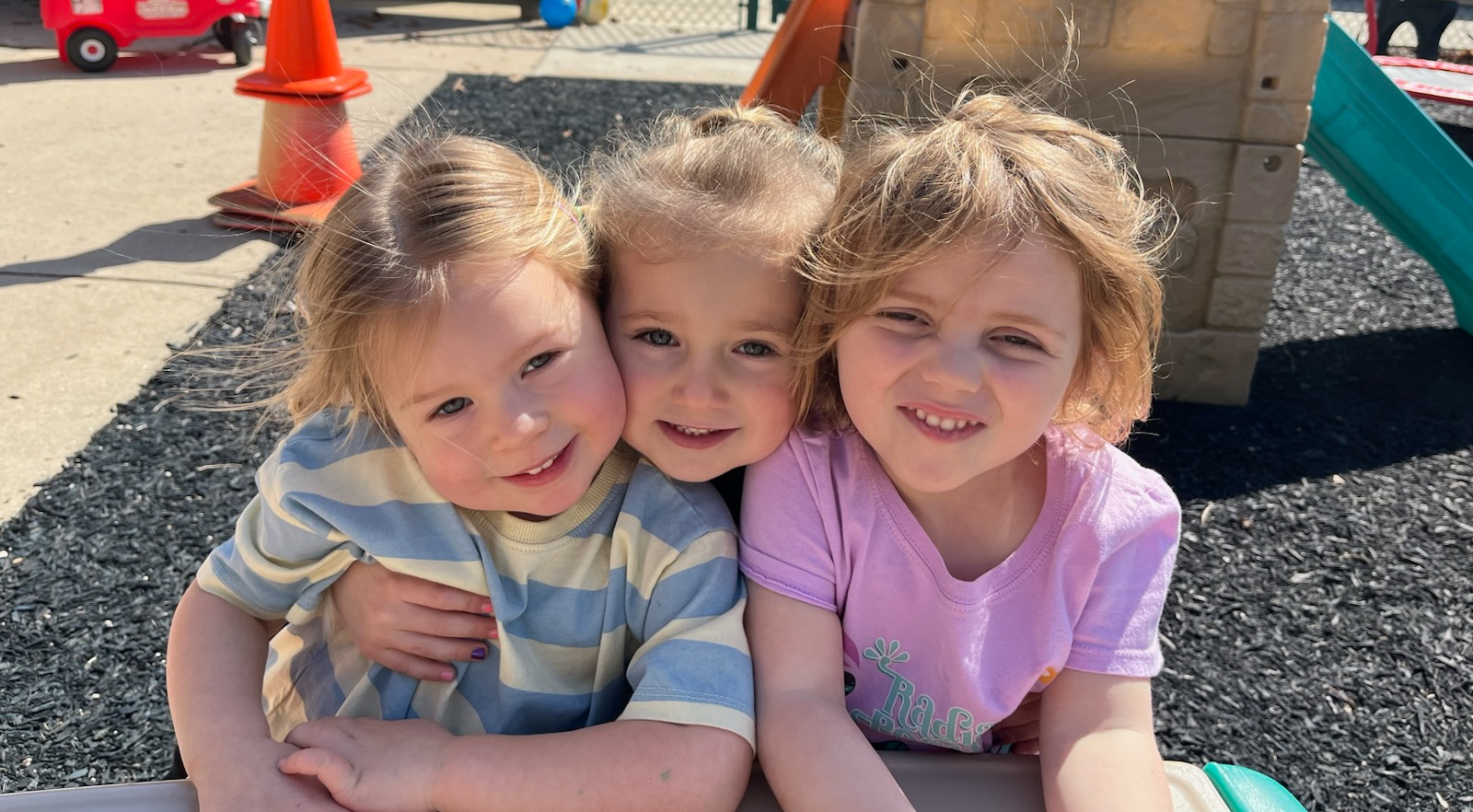 Three children smiling and embracing at an outdoor playground with a toy slide and traffic cones in the background, under a sunny sky.