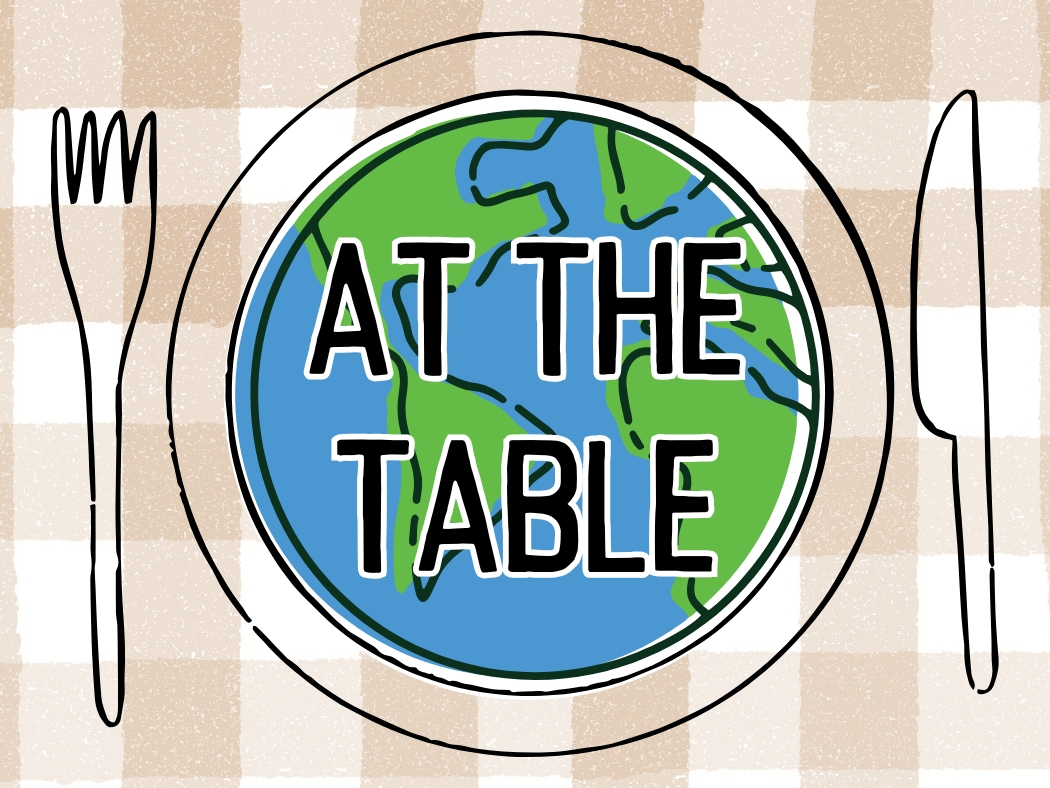 A plate with a globe design, labeled "At The Table," sits on a checkered tablecloth with fork and knife.