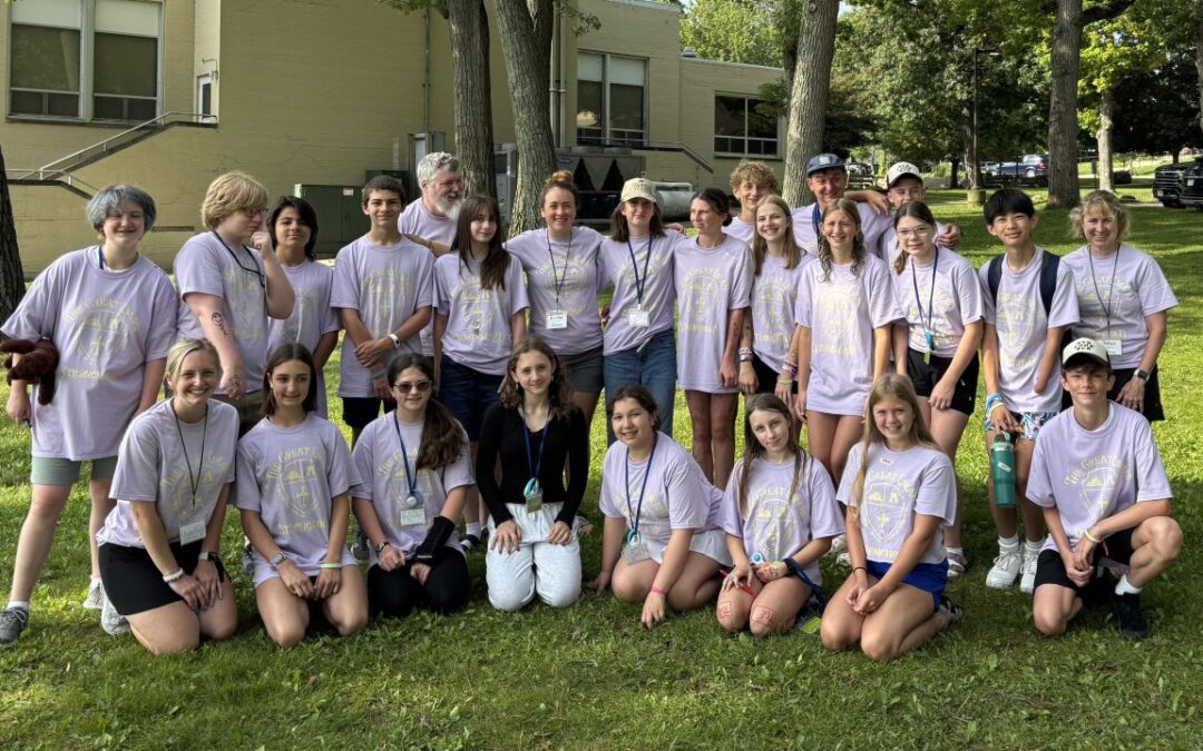 A group of people and children in matching shirts pose on grass in front of a building, surrounded by trees.