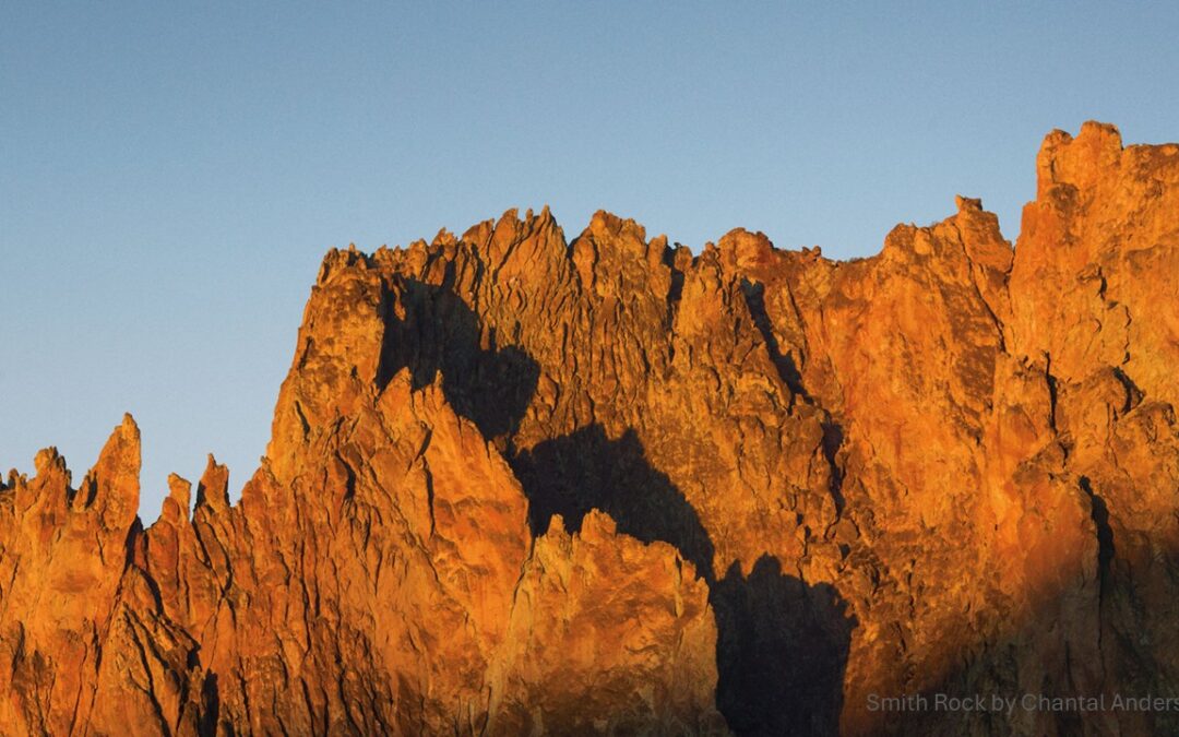 Smith Rock's towering, sunlit cliffs glow orange and red under a clear blue sky, showcasing dramatic, rugged rock formations.