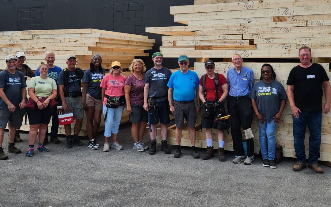 A group of people stands in front of stacked lumber, possibly participating in a community building project.